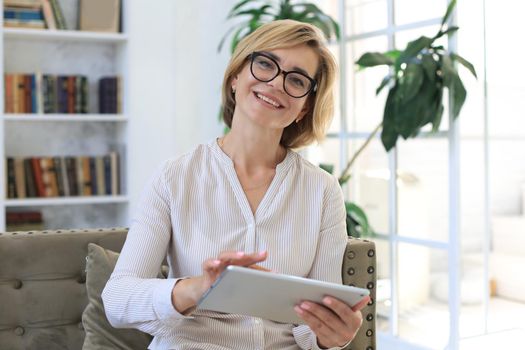 Cheerful middle aged woman sitting on sofa, using computer tablet apps, looking at screen,reading good news in social network, shopping or chatting online