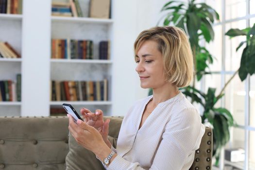 Smiling middle aged woman holding phone, using mobile device apps, looking at screen, while sitting on couch