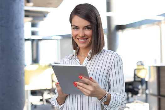 Portrait of young businesswoman looking at camera, holding touch pad while standing in modern office space interior