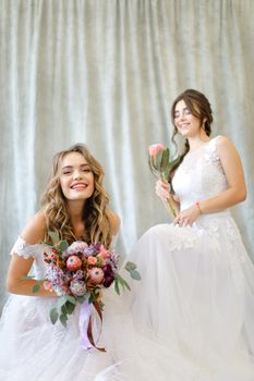 Two caucasian brides wearing white dress and sitting at studio with flowers. Concepr of bridal photo session and wedding.