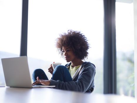 Young african american woman smiling sitting near bright window while looking at open laptop computer on table and holding white mug in her luxury home