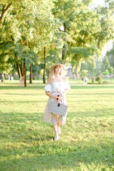 Young pretty girl walking in garden and keeping bag, sunglasses and hat. Concept of walking in park and summer fashion garb.