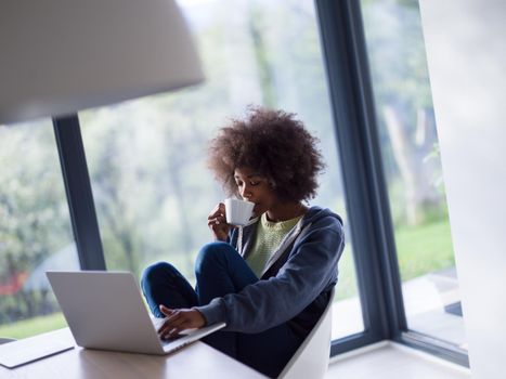 Young african american woman smiling sitting near bright window while looking at open laptop computer on table and holding white mug in her luxury home