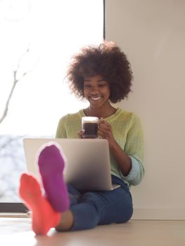 Young african american woman smiling sitting on the floor near bright window while looking at open laptop computer and holding mug at home