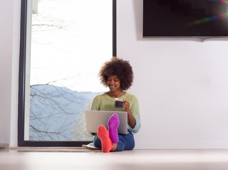 Young african american woman smiling sitting on the floor near bright window while looking at open laptop computer and holding mug at home