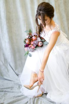 Young caucasian bride with bouquet of flowers putting on shoes at studio. Concept of bridal photo session and wedding.