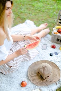 Young blonde woman sitting on plaid near fruits and hat, eating watermelon, grass in background. Concept of summer picnic and resting on weekends in open air.