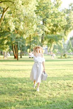 Young nice girl walking in garden and keeping bag, sunglasses and hat. Concept of walking in park and summer fashion.