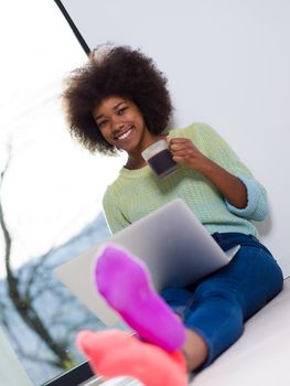 Young african american woman smiling sitting on the floor near bright window while looking at open laptop computer and holding mug at home