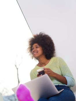 Young african american woman smiling sitting on the floor near bright window while looking at open laptop computer and holding mug at home