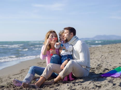 Family with little daughter resting and having fun making soap bubble at beach during autumn day