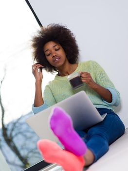 Young african american woman smiling sitting on the floor near bright window while looking at open laptop computer and holding mug at home