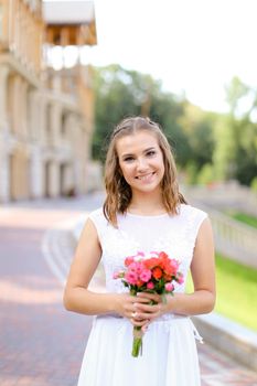 Young bride wearing white dress and standing outside with flowers. Concept of wedding and bridal photo session, floristic art.