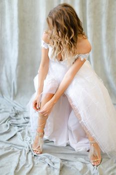 Young blonde bride in white dress putting on shoes at studio. Concept of bridal photo session and wedding.
