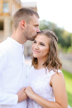 Caucasian groom kissing bride and holding hands outdoors. Concept of wedding and bridal photo session, relationship.