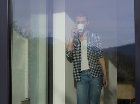 beautiful young woman drinking morning coffee by the window in her home