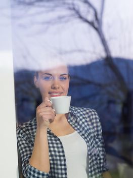 beautiful young woman drinking morning coffee by the window in her home