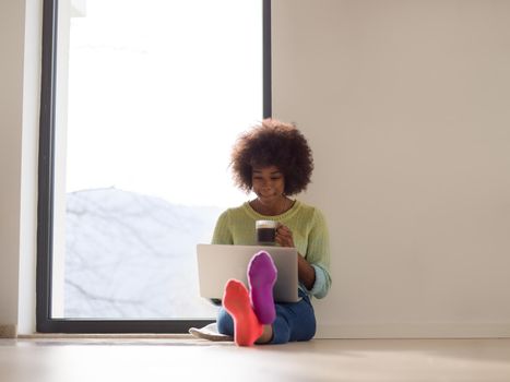 Young african american woman smiling sitting on the floor near bright window while looking at open laptop computer and holding mug at home