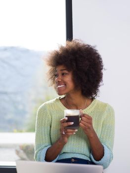 Young african american woman smiling sitting on the floor near bright window while looking at open laptop computer and holding mug at home