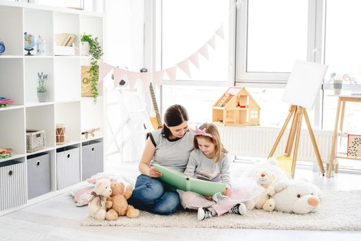 Beautiful mother and daughter reading book sitting on floor in kids room