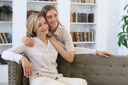 Beautiful middle aged mom and her adult daughter are hugging and smiling while sitting on couch at home