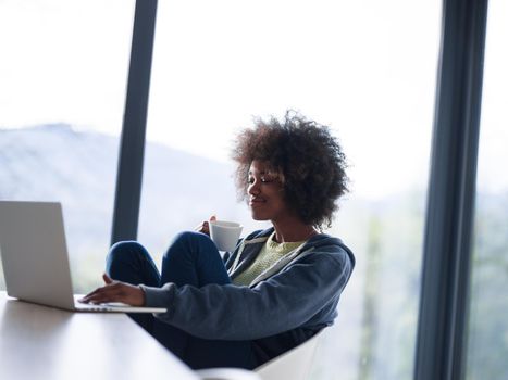 Young african american woman smiling sitting near bright window while looking at open laptop computer on table and holding white mug in her luxury home