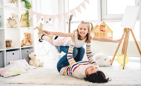 Laughing daughter flying on mother's legs in bright room