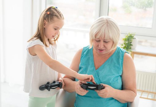 Little girl teaching elderly woman to play on joystick indoors