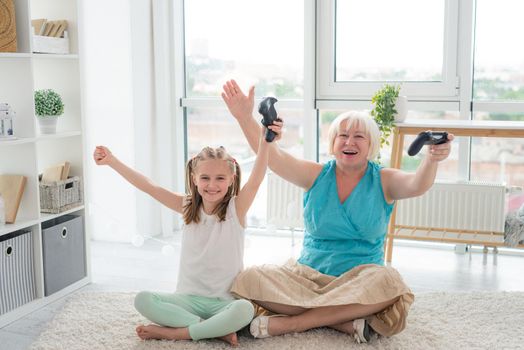 Happy grandma and cute granndaughter raising hands with joysticks up sitting on floor in light room