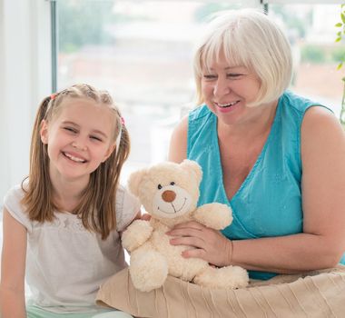 Happy grandmother and granddaughter playing with plush teddy indoors
