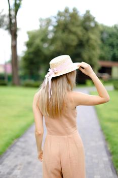 Back view of young blonde female person in body color overalls and hat standing in yeard. Concept of fashion and summer season.