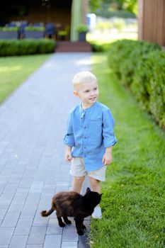 Little blond boy in jeans shirt standing with cat in yard. Concept of kids, childhood and pets.