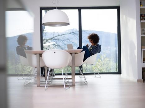 Young african american woman smiling sitting near bright window while looking at open laptop computer on table and holding white mug in her luxury home