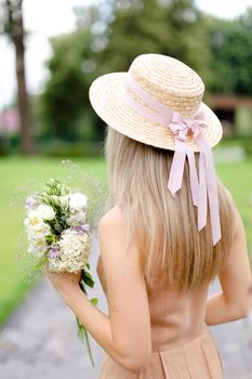 Back view of young blonde girl in body color overalls and hat standing in yeard with flowers. Concept of fashion and summer season.