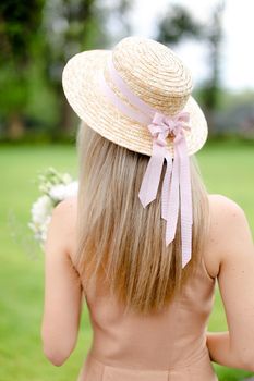 Back view of young female person in body color overalls and hat standing in yeard. Concept of fashion and summer season.