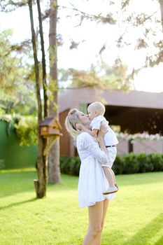 Young blonde mom walking with child on grass in yard. Concept of motherhood and kids, summer season.