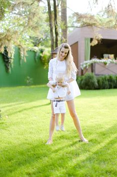 Young happy mom walking with child on grass in yard. Concept of motherhood and kids, summer season.