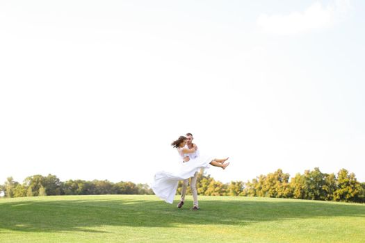 Happy groom holding bride on grass in white sky background. Concept of wedding photo session on nature.