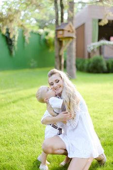 Young caucasian mother playing and sitting with little kid on grass in yard. Concept of motherhood and child.