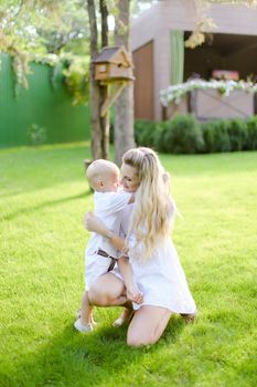 Young blonde mother playing and sitting with little kid on grass in yard. Concept of motherhood and child.