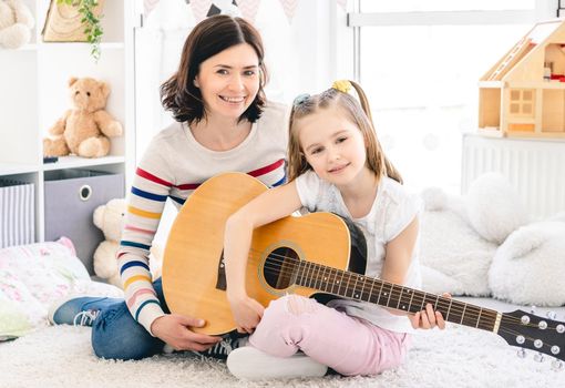 Happy mother with cute daughter playing guitar at home