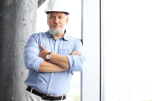 Thoughtful mature architect wearing hardhat and looking away while standing near the window in new modern building