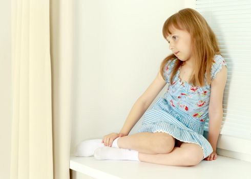 A nice little girl is sitting by the window with blinds. The concept of a happy childhood, construction, production of horizontal blinds.