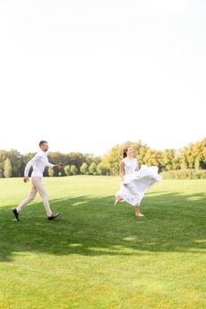 Groom and bride in white dress running and playing on grass. Concept of wedding photo session on open air and nature.