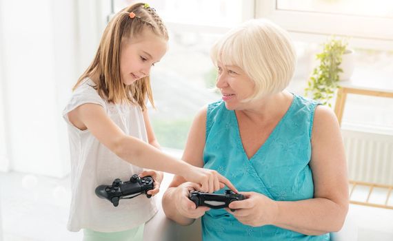 Little girl teaching elderly woman to play on joystick indoors