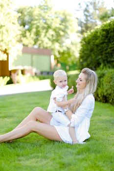 Young pretty mother sitting with little child on grass in yard. Concept of motherhood and summer vacations.