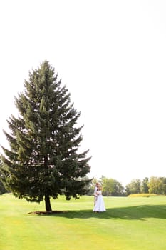 Young european couple walking near green big spruce on grass in white sky background. Concept of evergreens and wedding.