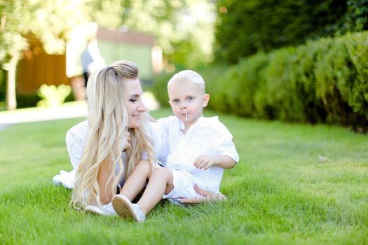 Young blonde mother sitting with little child on grass in yard. Concept of motherhood and summer vacations.