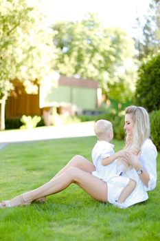 Young happy mother sitting with little child on grass in yard. Concept of motherhood and summer vacations.