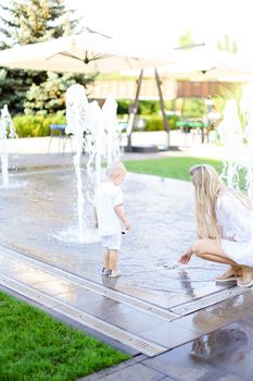 Young mother playing with little male baby outside near fountain. Concept of walking in city and motherhood.